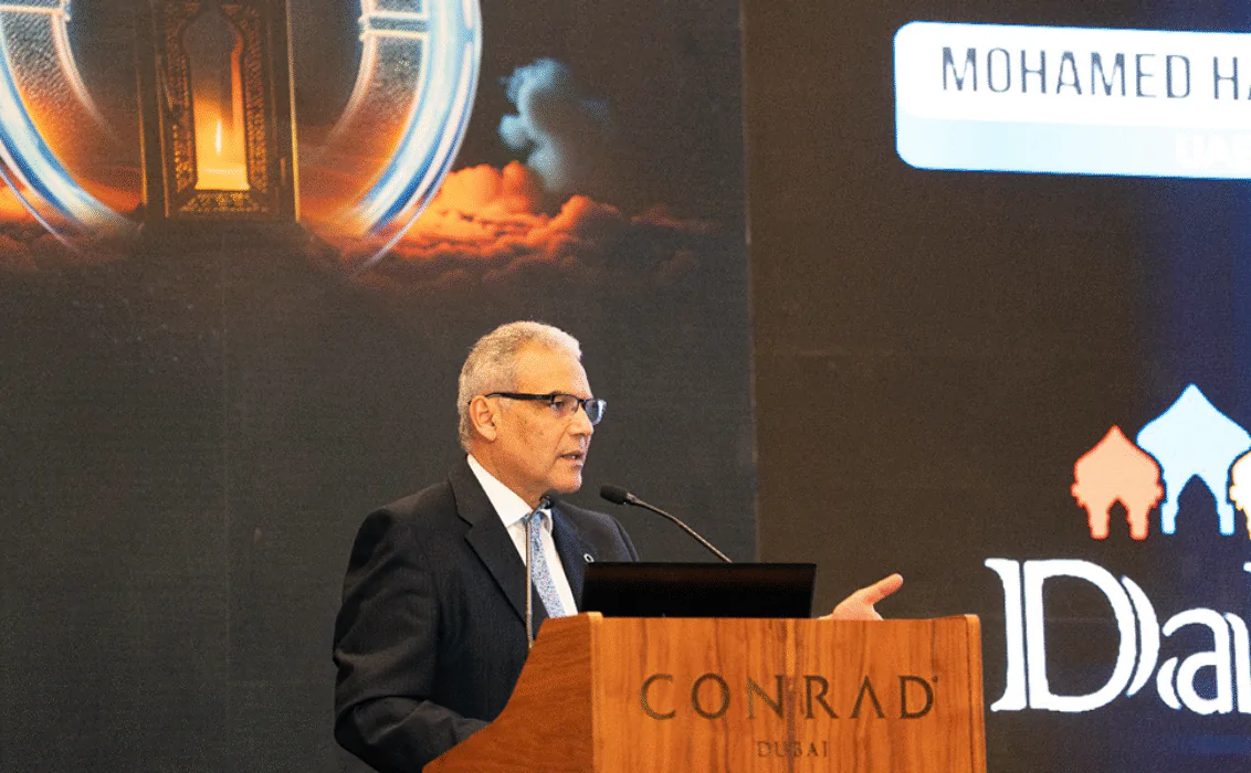 Prof Mohamed Hassanein stands at a podium. He is wearing a dark suit and tie and speaks into a microphone while gesturing with one hand. Behind him, a large event screen displays vibrant graphics, including an illuminated lantern motif and text elements, alongside the logo of the DaR programme.